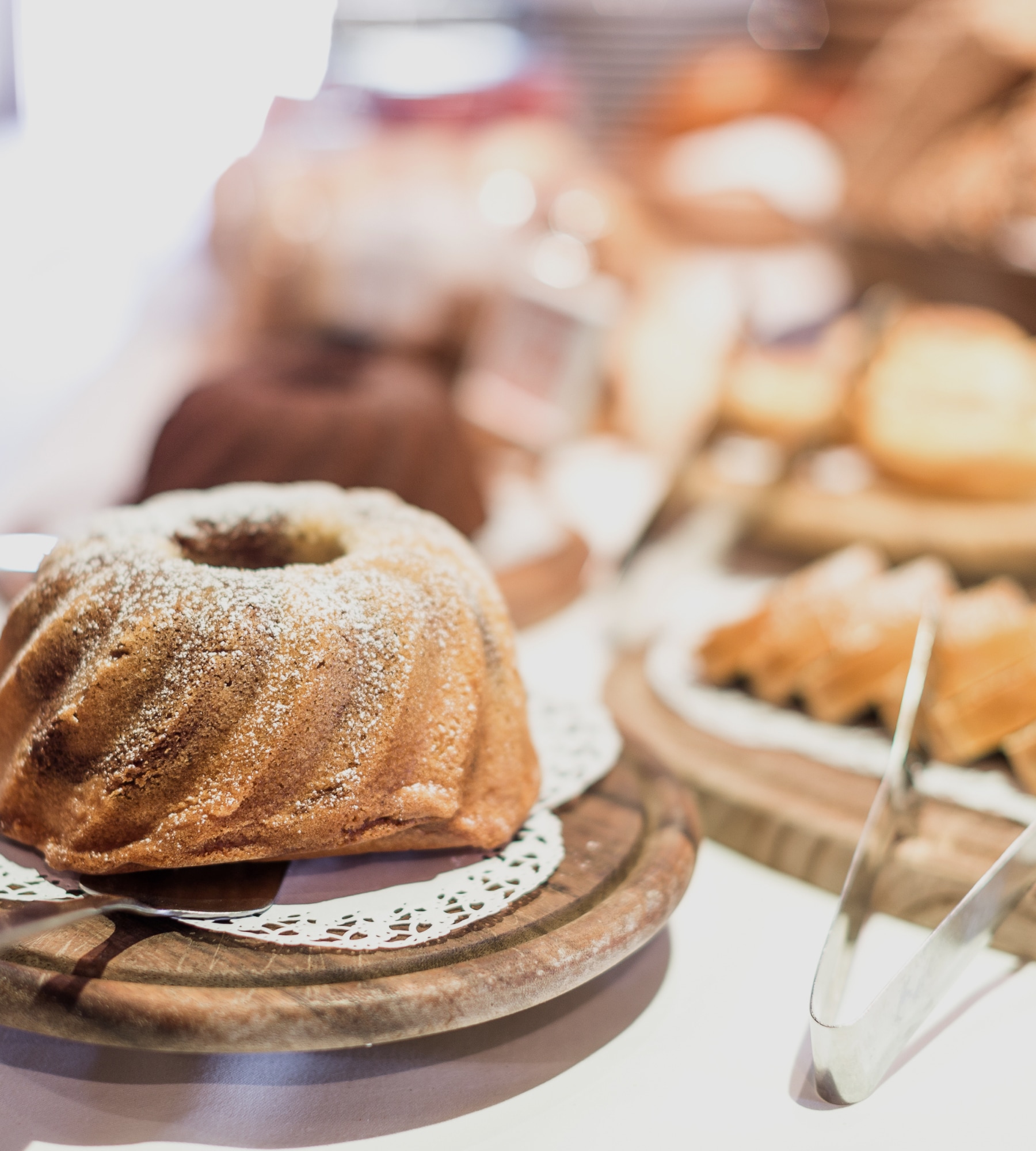 Ein Gugelhupf mit Puderzucker auf einem Holzbrett, im Hintergrund weiteres Gebäck auf einem Buffet.
