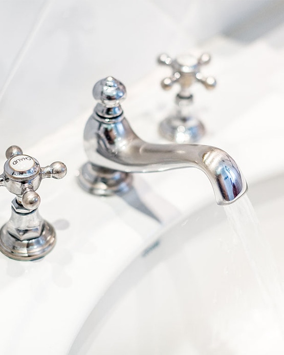 Klassischer, verchromter Wasserhahn mit Kreuzgriffen, aus dem Wasser in ein weißes Waschbecken fließt.