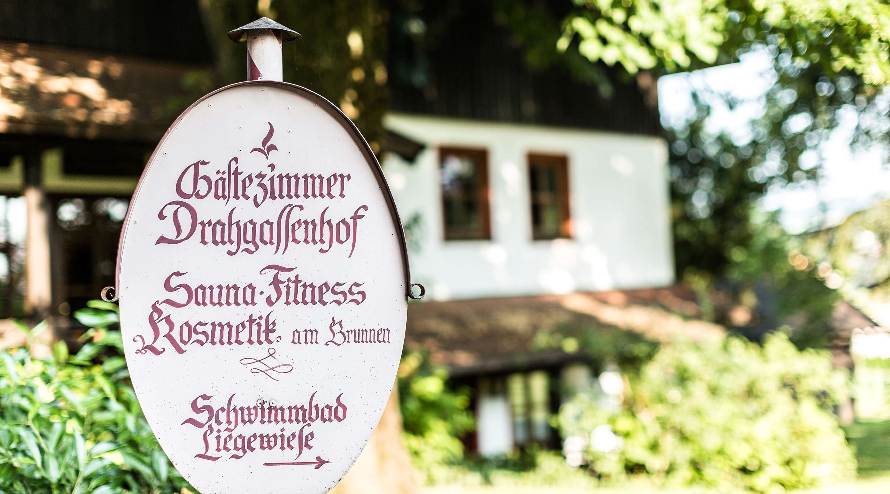 Schild „Gästezimmer Drahgassenhof“ mit Infos zu Sauna, Fitness und Schwimmbad vor einem ländlichen Gebäude.