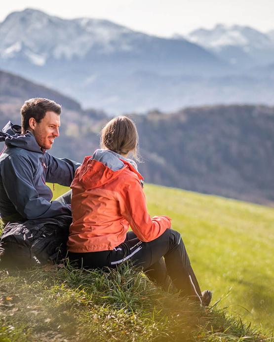 Ein Paar in Outdoor-Kleidung rastet auf einer Bergwiese und genießt den Blick auf die Berge.