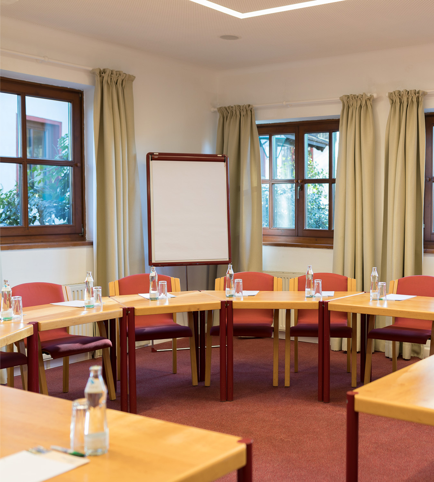 Ein Tagungsraum im Kreis aufgestellt mit Holztischen, roten Stühlen, Flipchart und Fenstern im Hintergrund.