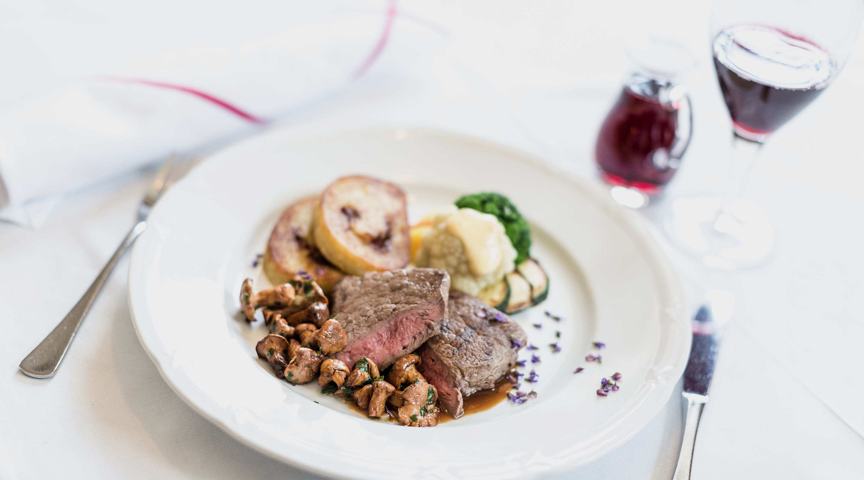 Plated venison steak with wild mushrooms, dumplings, vegetables, and a glass of red wine.