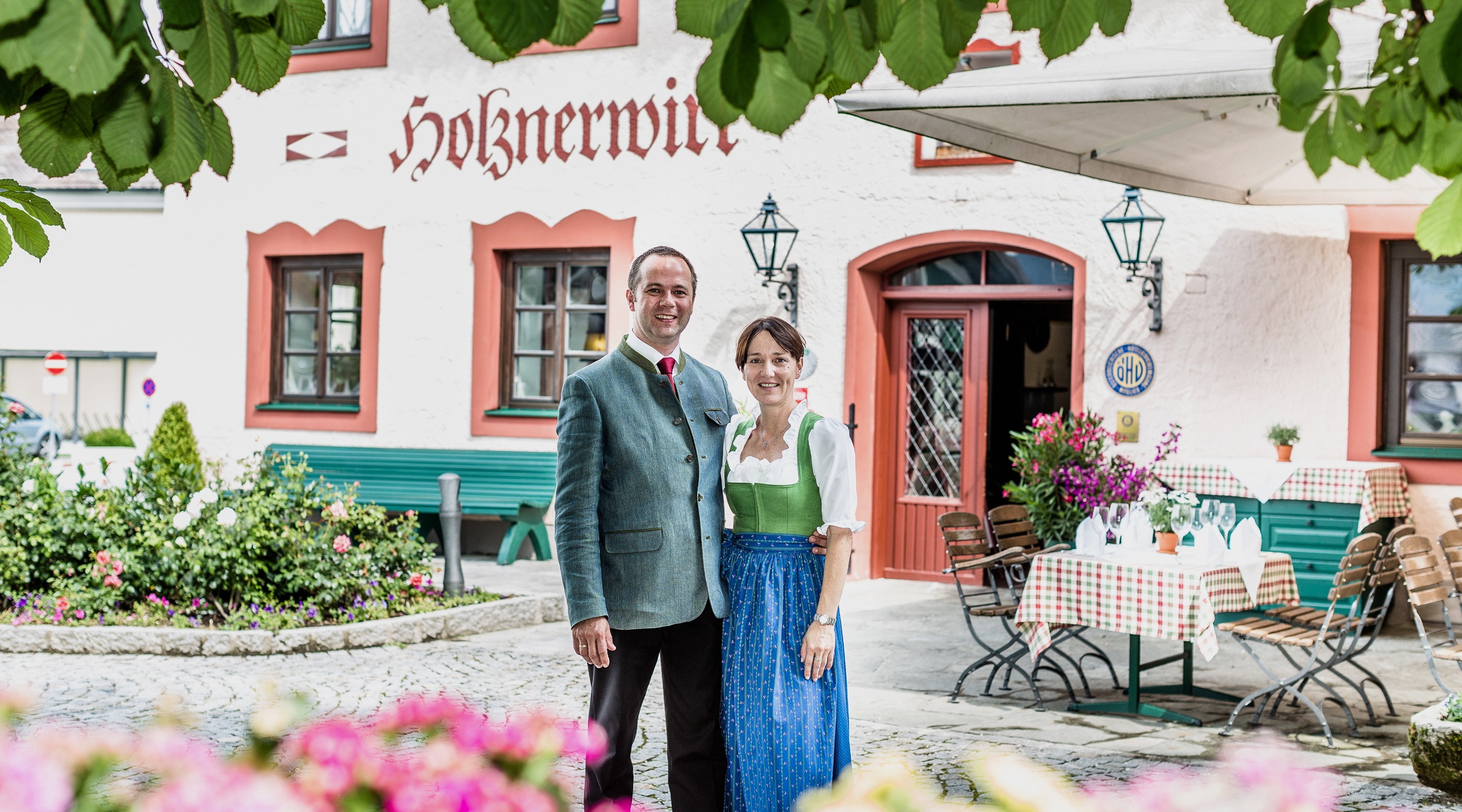 A man and woman in traditional Austrian attire stand smiling in front of the Holznerwirt hotel.