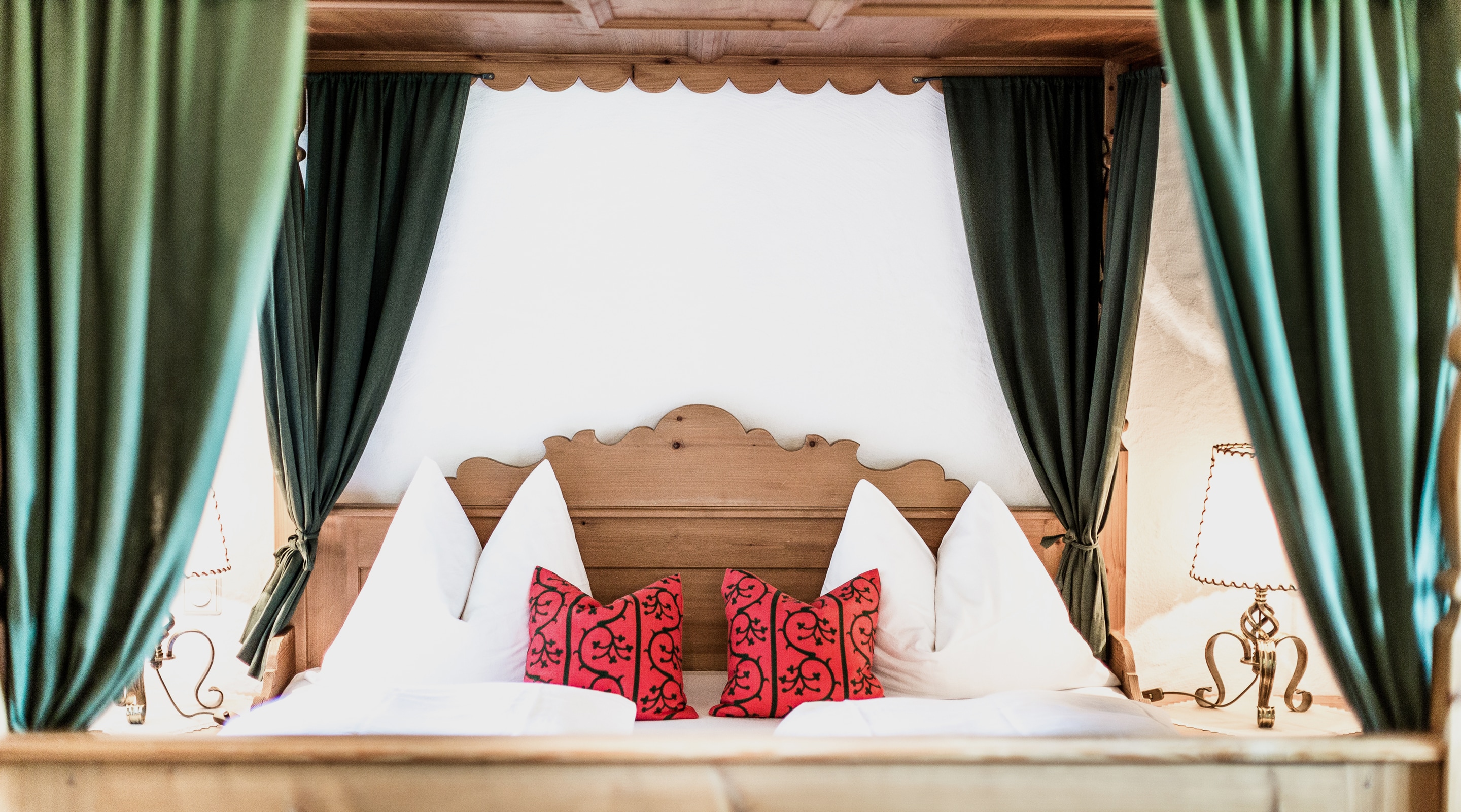 Rustic four-poster wooden bed with white pillows, red patterned cushions, and dark green curtains.
