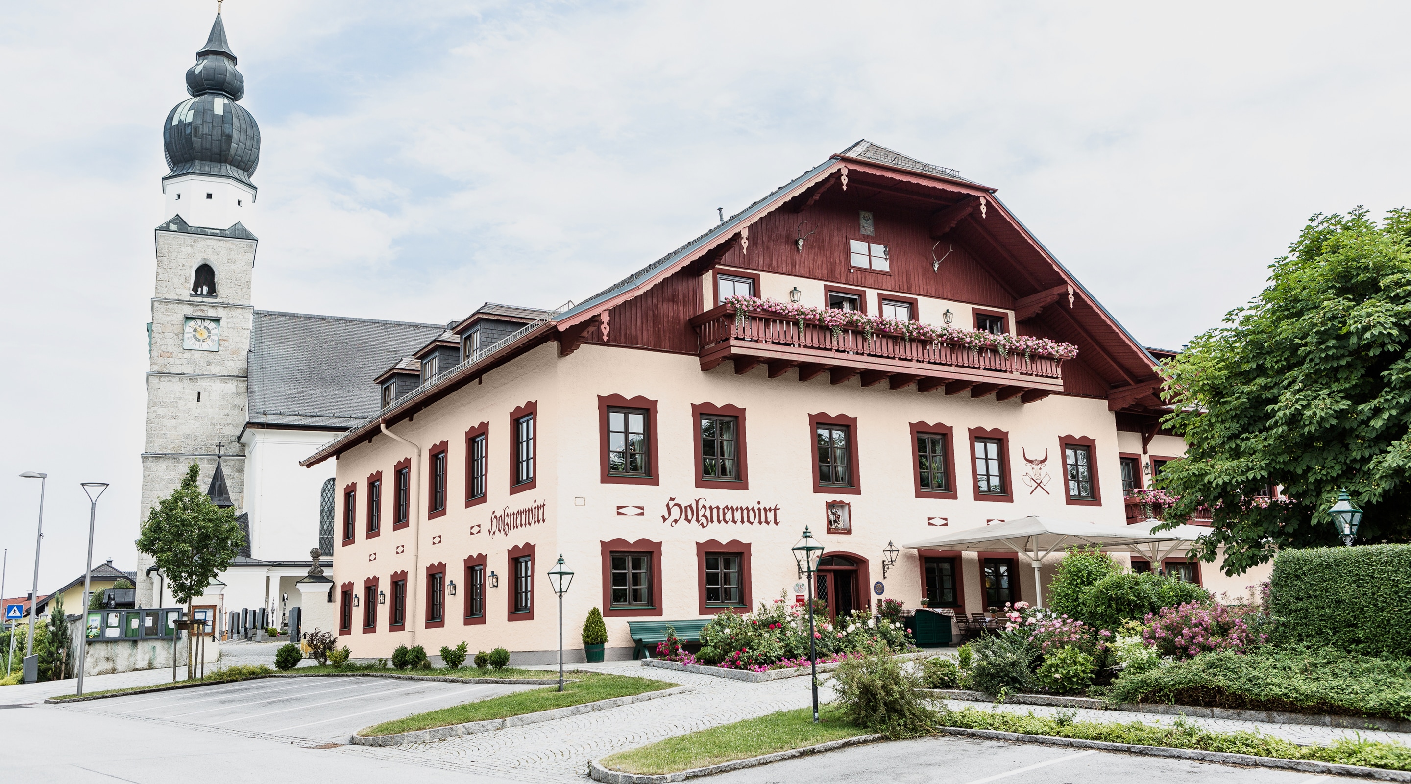 Der Gasthof Holznerwirt in Eugendorf mit dem Kirchturm im Hintergrund an einem bewölkten Tag.