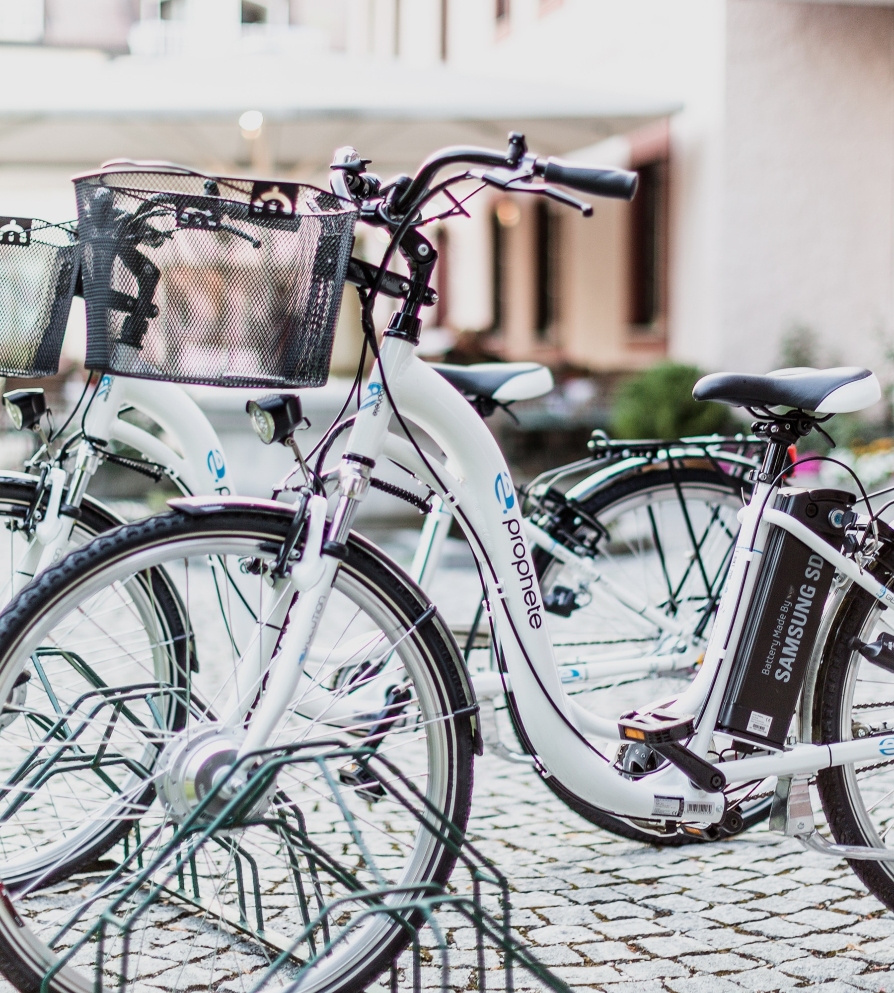 White Prophete e-bikes with front baskets parked on a cobblestone street, showing Samsung SDI batteries.