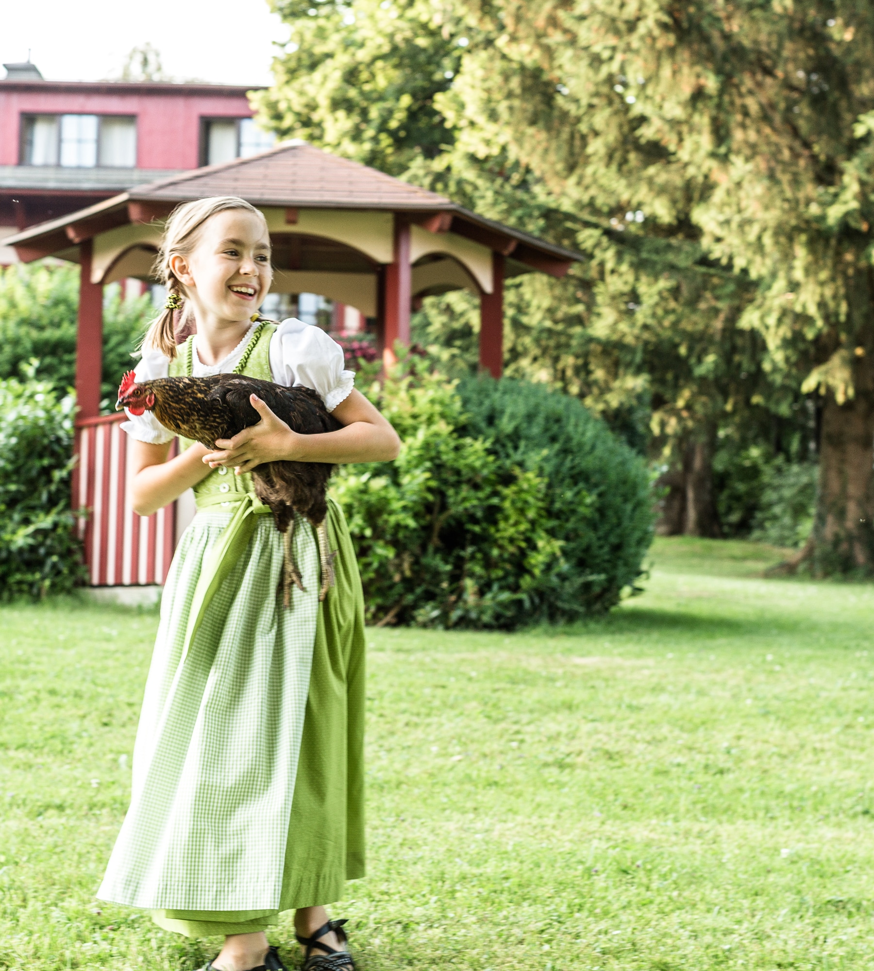 A smiling girl in a green dirndl holds a chicken in a grassy garden, with a red gazebo in the background.