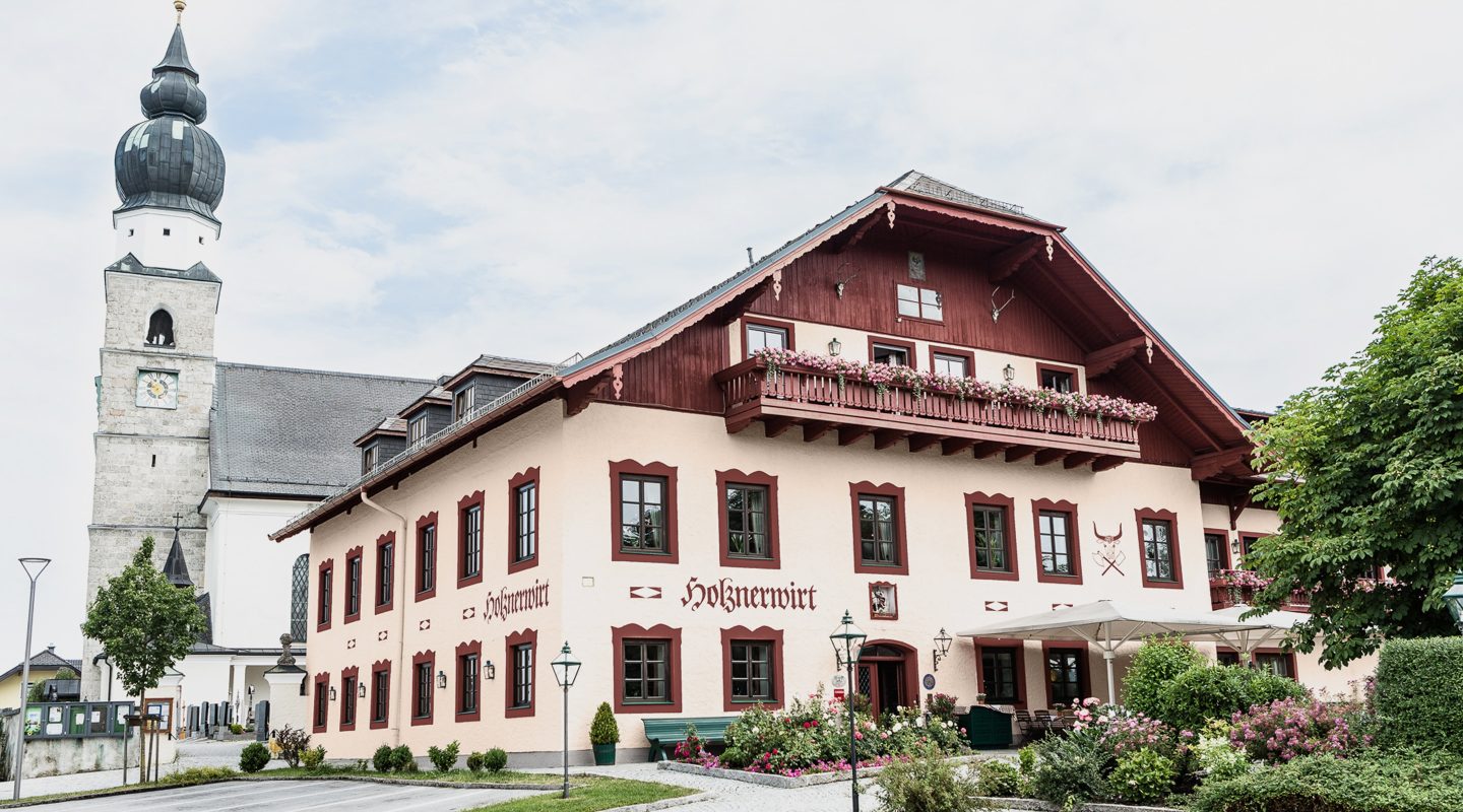 Der Gasthof Holznerwirt mit blumengeschmücktem Balkon neben einer Kirche mit Zwiebelturm.