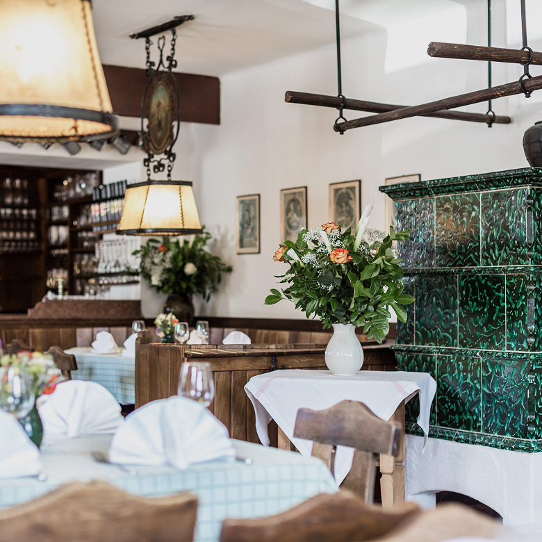 Rustikales Restaurant mit gedeckten Tischen, Blumenstrauß und einem grünen Kachelofen im Hintergrund.