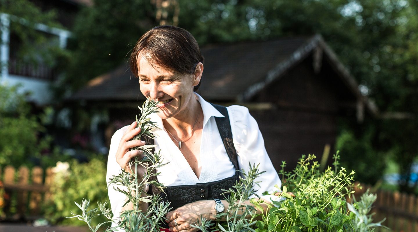 Eine lächelnde Frau in Tracht riecht an frischen Kräutern im Garten vor einer Holzhütte.