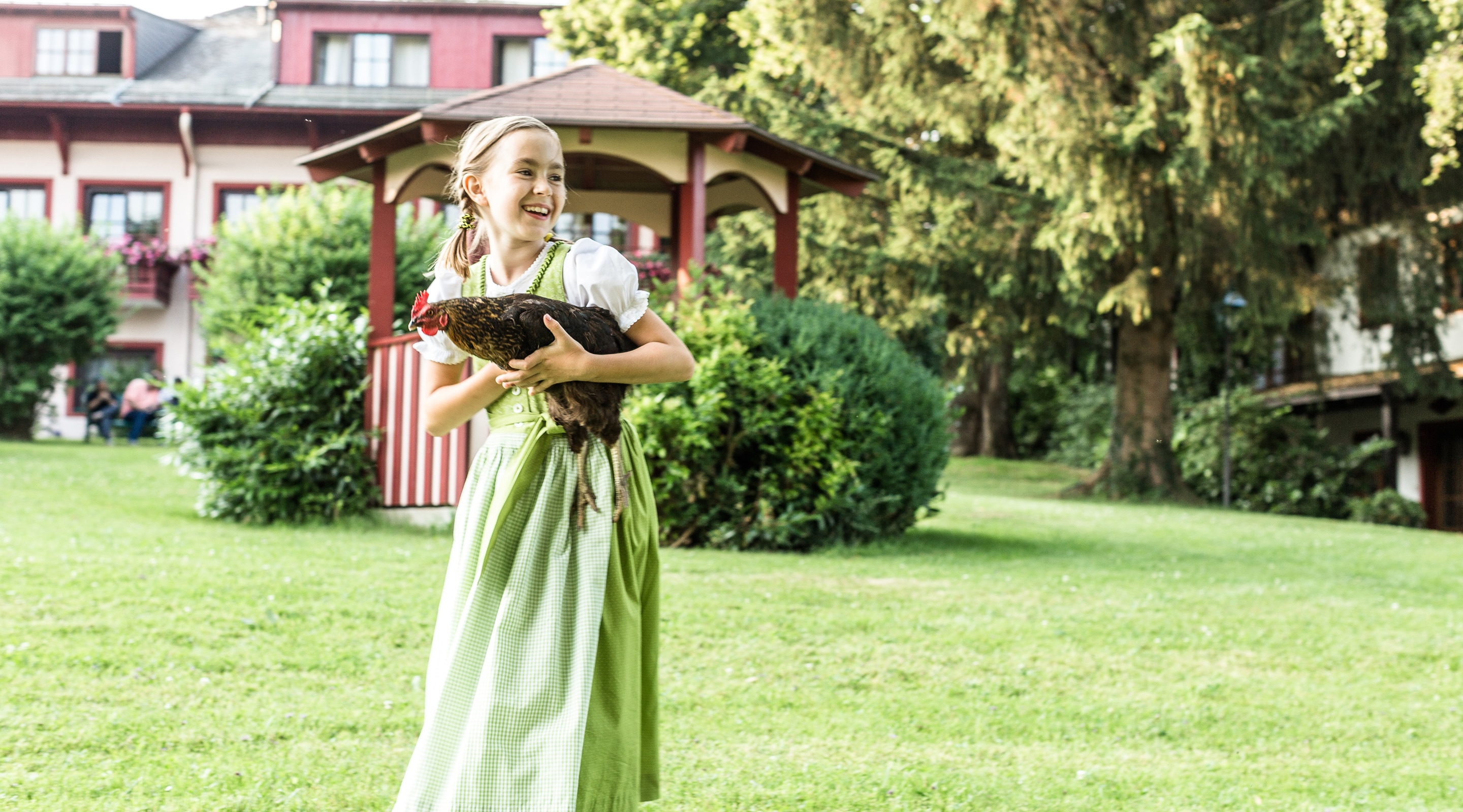Ein lachendes Mädchen im grünen Dirndl hält ein braunes Huhn im Arm auf einer grünen Wiese vor einem Pavillon.