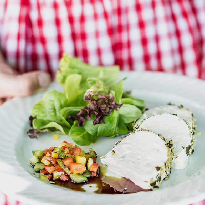 Ziegenkäserolle in Kräutermantel mit buntem Gemüsetatar, Salat und Balsamico auf einem weißen Teller serviert.