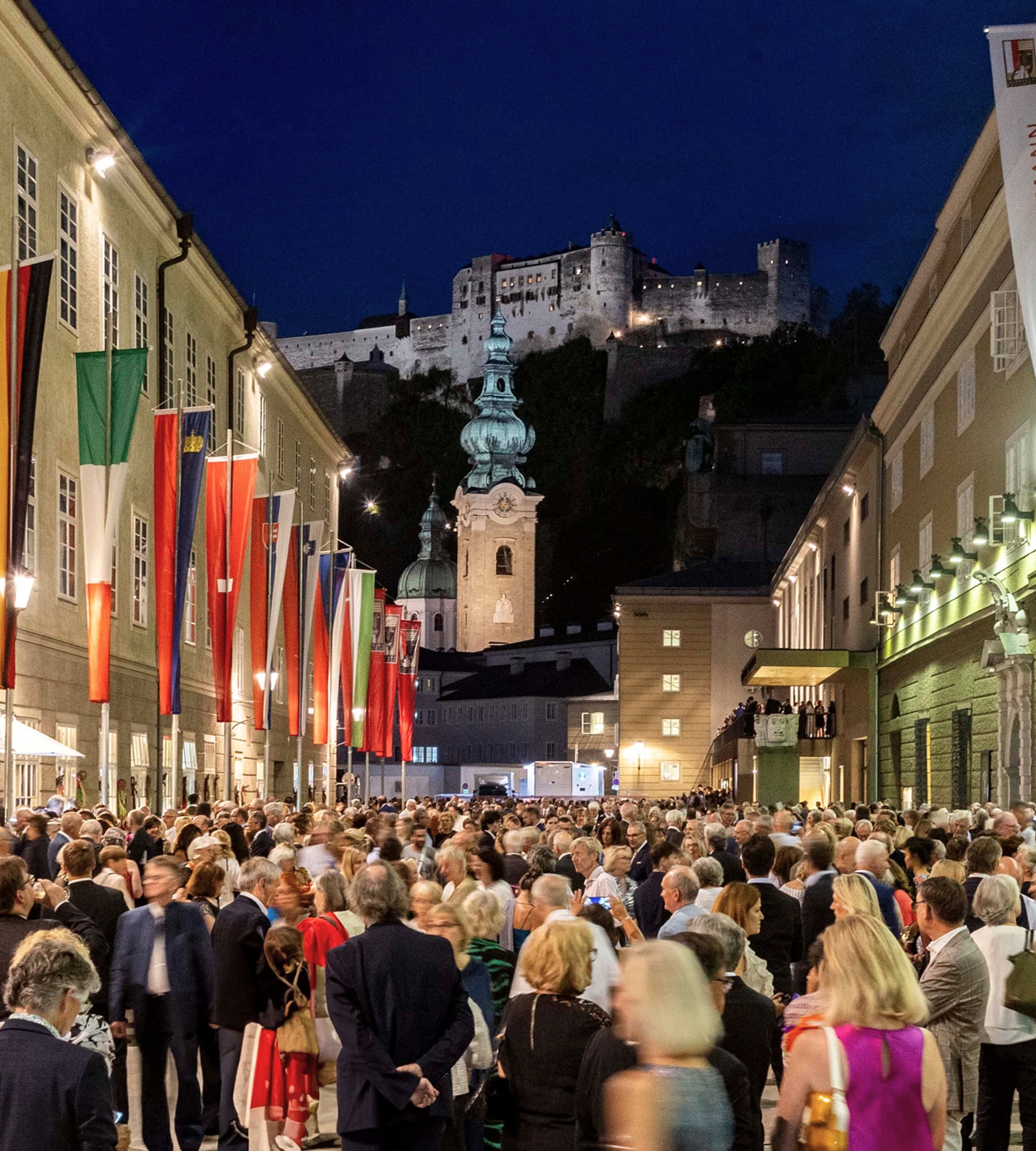 Menschenmenge in einer Gasse in Salzburg bei Nacht mit Blick auf die beleuchtete Festung Hohensalzburg.