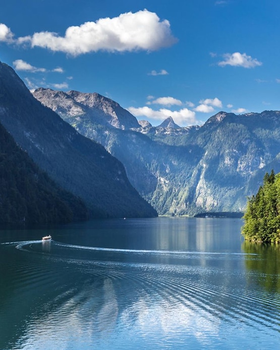 Ein Boot fährt auf dem blauen Königssee, umgeben von hohen Bergen unter einem leicht bewölkten Himmel.