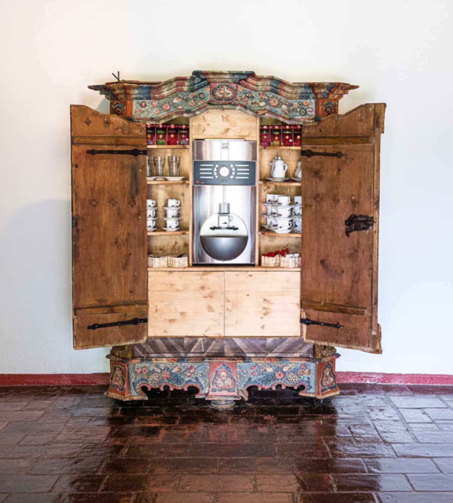 Ein antiker, bemalter Holzschrank, in dem sich eine moderne Kaffeemaschine und Geschirr befinden.